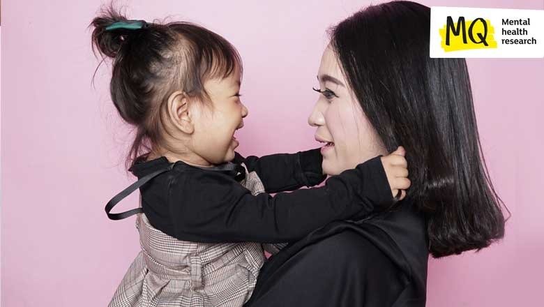 Against a pink background a young woman is holding a happy little girl who is smiling with her arms around the woman's neck