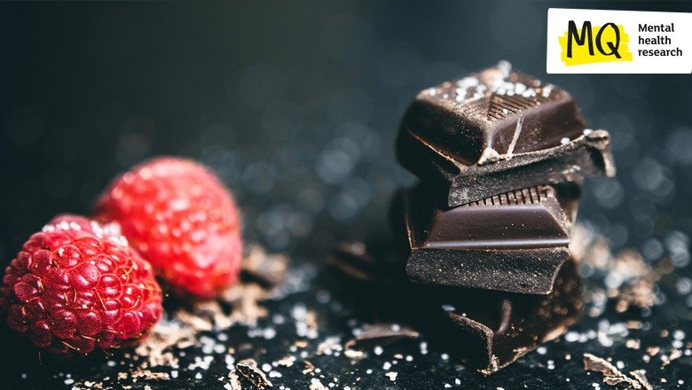 2 raspberries sit next to 3 squares of very dark chocolate with chocolate shavings surrounding them atop a black slate surface