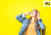 Against a yellow background a woman in a denim jacket and orange T shirt smiles upwards wearing yellow sunglasses and holding her hands to her ears as though she is listening to music on headphones