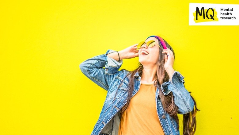 Against a yellow background a woman in a denim jacket and orange T shirt smiles upwards wearing yellow sunglasses and holding her hands to her ears as though she is listening to music on headphones