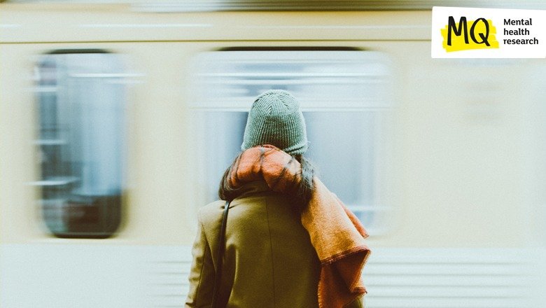 A person stands with back to camera wearing a coat, hat and scarf facing a train carriage ahead of them that is blurred perhaps because it is pulling into the station