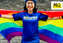 Amazin LeThi grins to camera with arms outstretched holding a Pride rainbow flag behind her. She stands in front of a brick wall wearing a blue t-shirt that reads "Athletes for Equality" and a beanie hat as well as rainbow wristbands