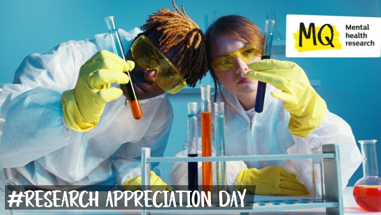 Against a vibrant blue background two researchers in white coats, yellow lab eye guards and yellow gloves each hold test tubes, one with orange liquid and one with dark purple liquid. There's a test tube rack with 4 further test tubes standing in it, holding orange, dark purple and light blue liquids in the foreground.