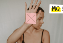 A women holds a hand out in front of her face which is turned away, as if to protect herself. On her hand is a pink post-it note with a large "X" written on it.