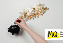 A black piggy bank sits on a blank grey background with coins laid upon it as if they are coming out of the piggy bank in a flurry. A human arm comes into frame to put a coin into the piggy bank