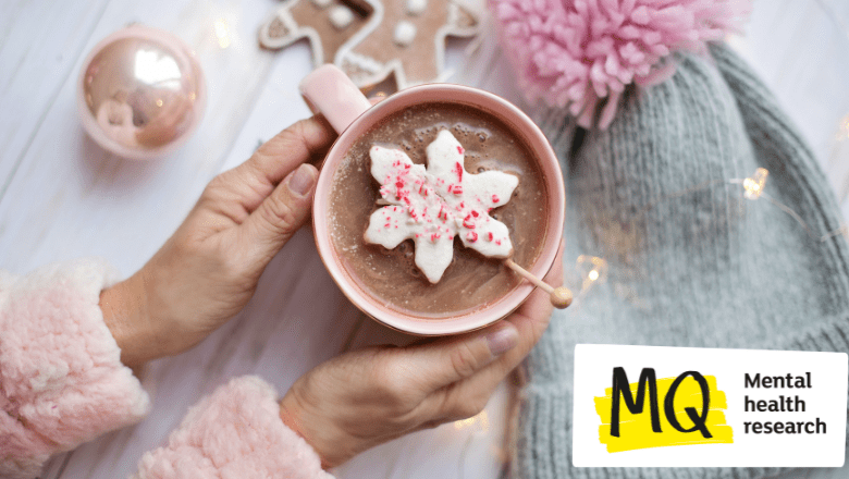 a mug of hot chocolate or cocoa is being held by a pair of hands that appear to belong to a female person. We can see her pink jumper sleeves as we look down from above onto the cocoa upon which there is a snowflake shaped decoration. Next to the mug is a pink bauble, two gingerbread men and a grey woollen hat with a pink bobble on.