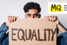 A male presenting figure holds a cardboard protest sign in front of his face, hiding his mouth and nose. The sign reads "Equality".
