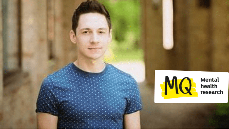 James Downs smiles to camera. He is white with short dark hair. He wears a blue T shirt with white dots. He stands in front of a sunlit building with an archway leading to out of focus trees.