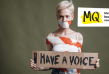 A woman with pixie cut blonde hair stands against a green background wearing jeans and a white strappy top holding a cardboard sign that says "I have a voice". She is tied up with hazard tape that binds her body and across her mouth.