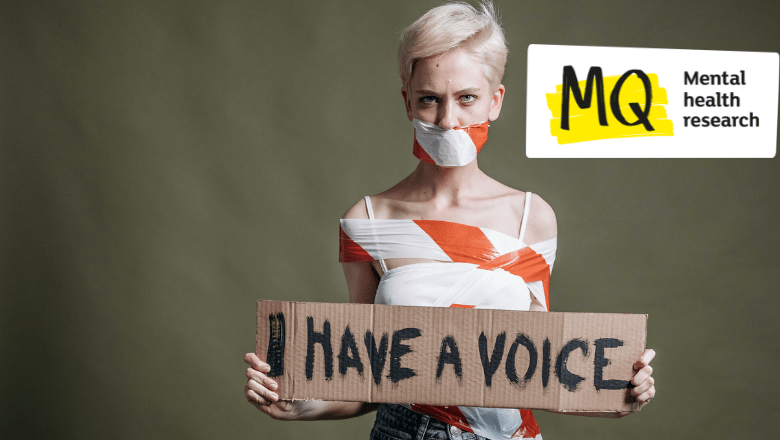 A woman with pixie cut blonde hair stands against a green background wearing jeans and a white strappy top holding a cardboard sign that says "I have a voice". She is tied up with hazard tape that binds her body and across her mouth.