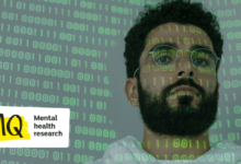 A man with a dark beard and glasses looks to camera. He is to the right of the image and we only see his face and neck. Projected onto him and a blank background is binary code in green.