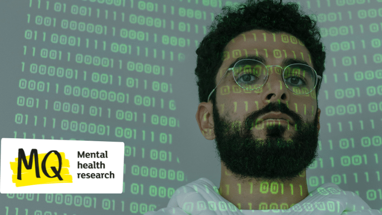 A man with a dark beard and glasses looks to camera. He is to the right of the image and we only see his face and neck. Projected onto him and a blank background is binary code in green.