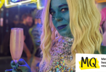 Painted blue, with blue make up, wearing a blonde wig and a glittery silver sequin dress, holding a gold champagne flute, a woman (Juliette Burton - the writer, cosplayer and comedian) smiles to camera.