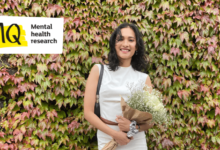Saira, the interviewee in this article, stands against a backdrop of green and pink vine leaves covering a wall. She has shoulder-length dark hair, white skin, is dressed in a sleeveless high neck white top and matching white skirt. On her right shoulder is a small brown handbag and cradled in her left arm is a bouquet of white and green gypsophila flowers wrapped in brown paper. She is smiling to camera warmly and relaxed.
