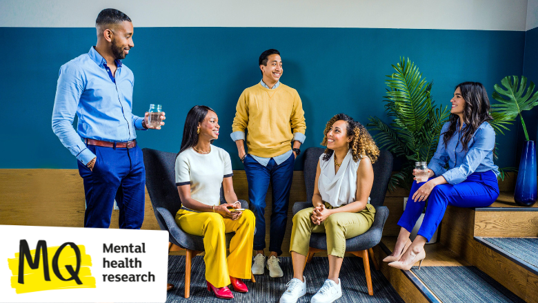 A group of people stand and sit against a blue wall. Some wear yellow and others wear blue. They are all different genders, races and ages.