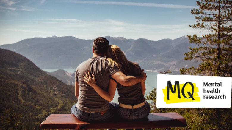 A couple sit with their backs to camera, arms around each other on a bench overlooking a beautiful view of a lake surrounded by forest-covered mountains with a blue sky and sunshine.