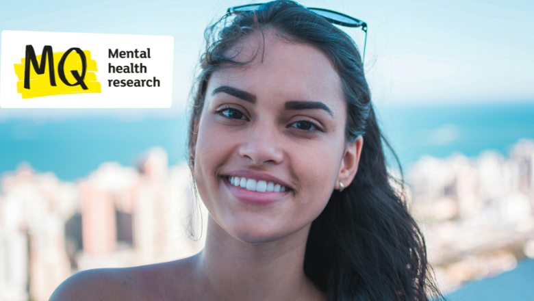 A woman with olive skin and dark hair smiles to camera in a headshot with what appears to be a sunny seaside town behind her out of focus.