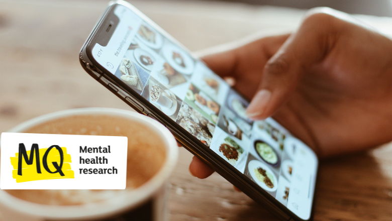 A close up of a smart phone held by a person's hand on a wooden table top with a grid of images on the screen, similar to an Instagram profile. In the foreground just out of shot is an open takeaway coffee cup.