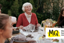 An older woman with white hair wearing a warm red cardigan and white roll neck top is laughing at a dinner table. Other women of various ages also sit around the dinner table engaged in conversation.