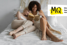 Two women snuggle up on a white bed together reading a book