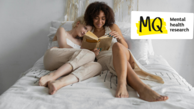 Two women snuggle up on a white bed together reading a book